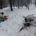 Kaksi sissiä sytyttää nuotiota märässä lumessa talvimetsässä.