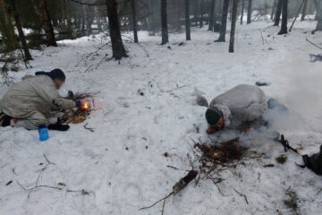 Kaksi sissiä sytyttää nuotiota märässä lumessa talvimetsässä.